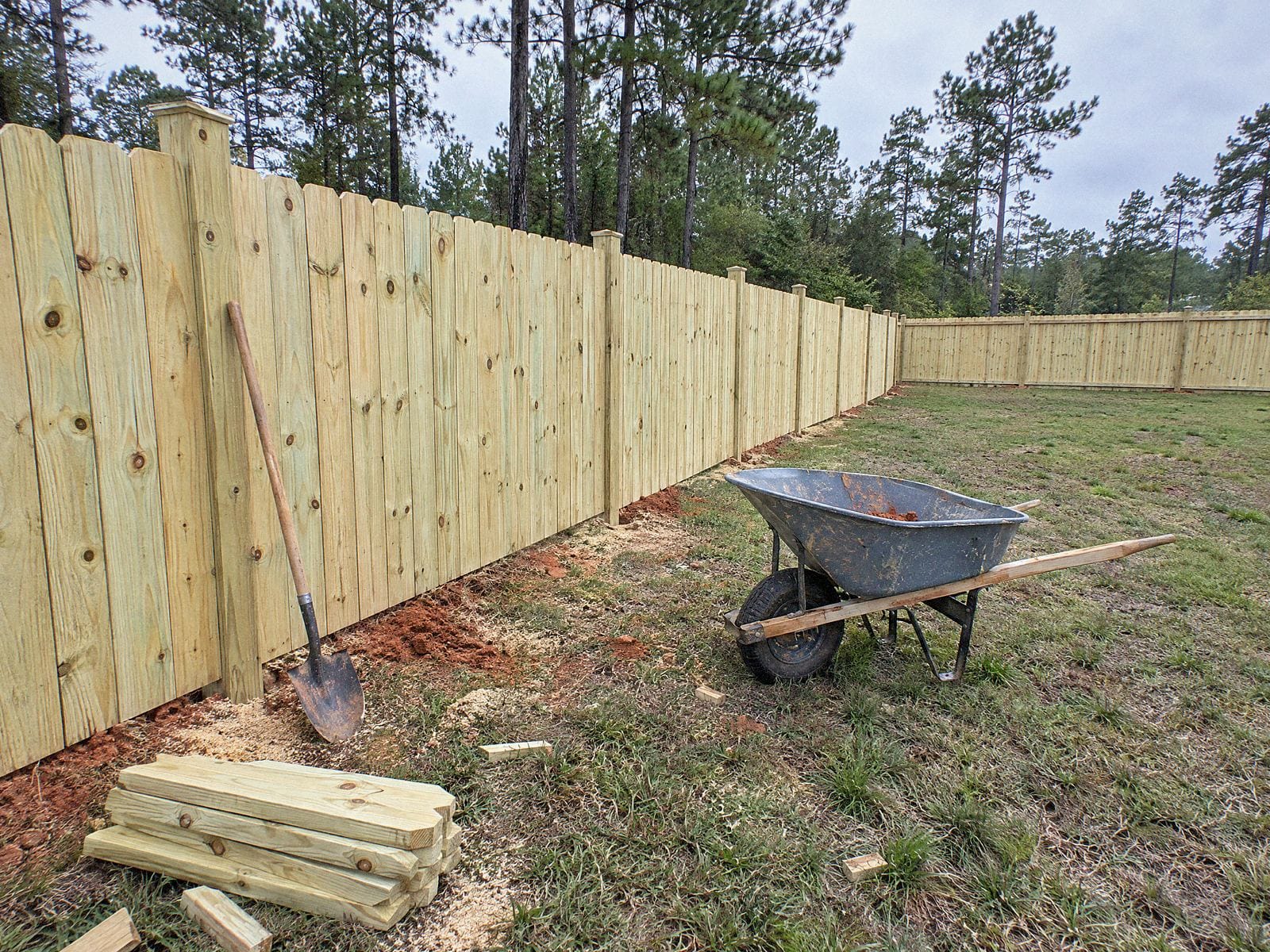Wood privacy fence line with a shovel leaning on a post, a wheelbarrow and a small stack of offcuts on the grass