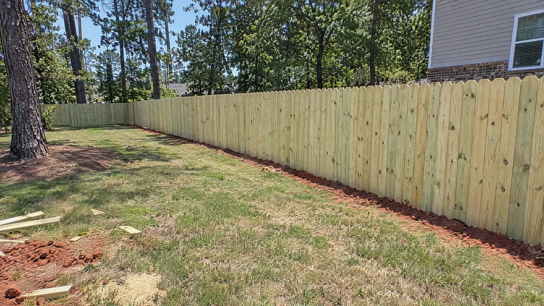 Long run of a six foot wood privacy fence with dog-ear pickets along a backyard lawn, tall pines behind it and red clay at its base